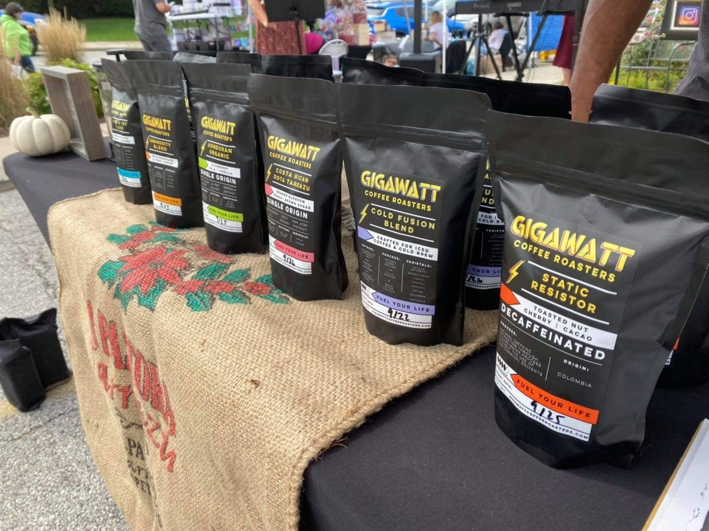 Gigawatt Coffee Roasters product lineup at a farmers market display, featuring blends like Costa Rican Single Origin, Cold Fusion Blend, and Static Resistor Decaf, arranged on a burlap-covered table."